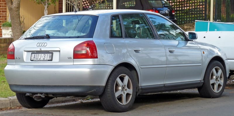 Traseira de um Audi A3 prata de cinco portas, da primeira geração, com lanternas vermelhas cortadas por uma faixa cinza-escura, os quatro anéis na tampa, a inscrição A3 à esquerda e 1.8 à direita