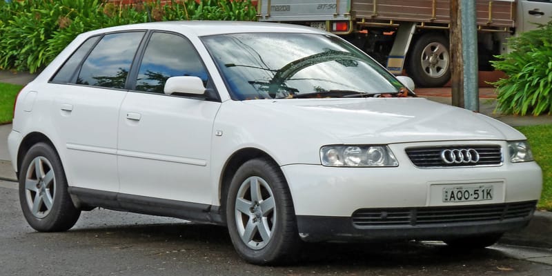Audi A3 branco de cinco portas, da primeira geração, estacionado no meio-fio, visto de frente e de lado, com faróis de lente lisa, grade com os quatro anéis e rodas de liga leve de cinco raios