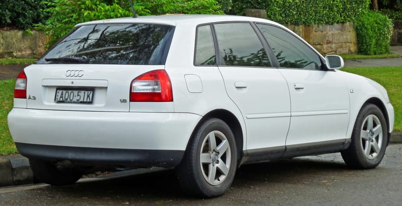 Traseira de um Audi A3 branco de cinco portas, da primeira geração, com lanternas vermelhas cortadas por uma faixa branca larga, os quatro anéis na tampa e as inscrições A3 e 1.8