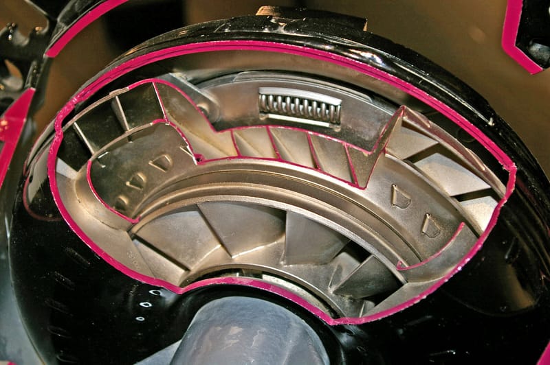 Conversor de torque em corte, com a carcaça preta aberta e bordas pintadas de magenta, deixando ver as aletas curvas de metal claro das rodas internas e uma mola espiral no alto