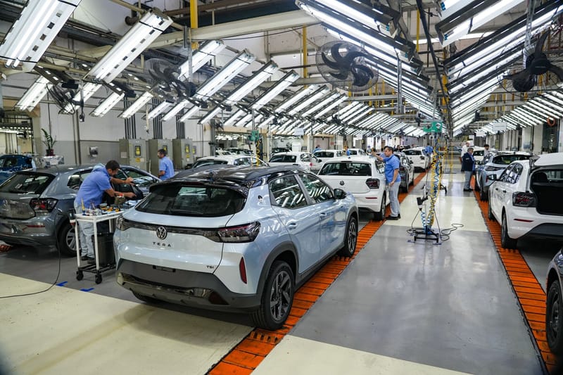 Interior de uma linha de montagem com teto de lâmpadas tubulares e ventiladores: em primeiro plano, um SUV Volkswagen Tera azul-claro com teto preto, visto de trás; à esquerda, um Polo cinza; à direita do Tera, um Polo branco; ao fundo, outros carros brancos, com funcionários de camisa azul trabalhando neles