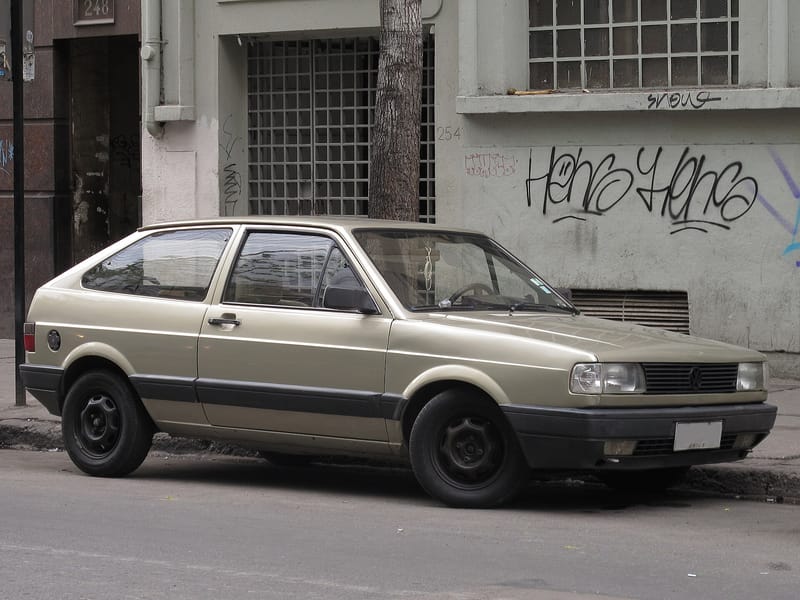 Volkswagen Gol de duas portas na cor bege, primeira geração, visto de lado e de frente, estacionado numa rua, com para-choques pretos e rodas de aço sem calota