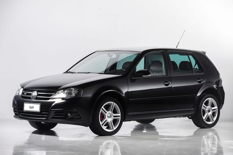 Golf GTI preto de quatro portas, da quarta geração reestilizada, fotografado em estúdio de fundo branco, visto de frente e de lado, com grade cromada, logotipo GTI na grade, rodas de liga leve de cinco raios duplos e pinças de freio vermelhas