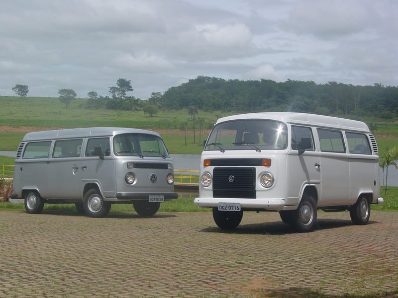 Duas Kombi de teto alto lado a lado num pátio de blocos de concreto: à esquerda uma prata, de frente lisa com uma faixa estreita de ventilação; à direita uma branca, com grande grade preta retangular entre os faróis