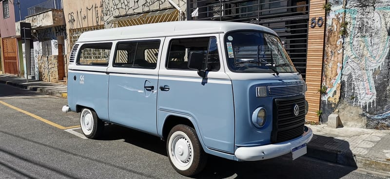 Kombi azul-clara com teto alto branco estacionada numa rua, vista de frente e de lado, com grade preta retangular entre os faróis redondos, para-choque branco e pneus de faixa branca