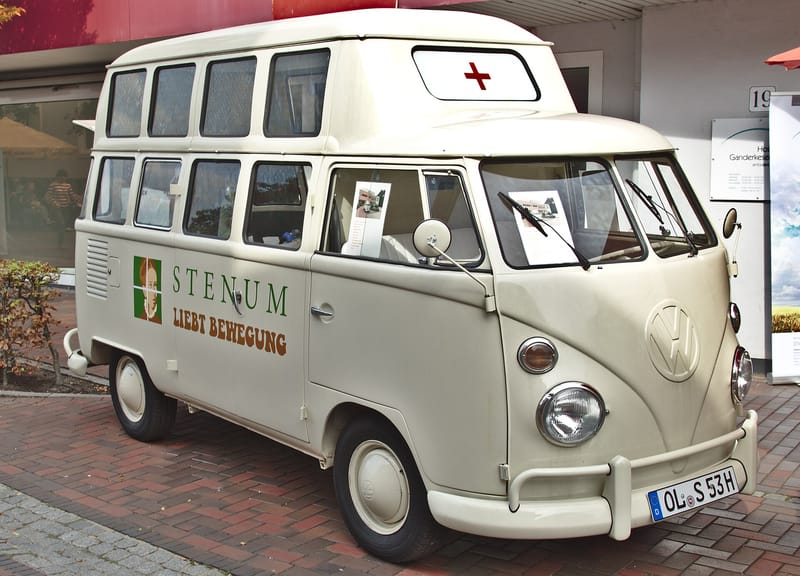 Kombi de primeira geração branca, com para-brisa dividido em duas peças, teto elevado com janelas e cruz vermelha na frente do teto, estacionada sobre piso de tijolos