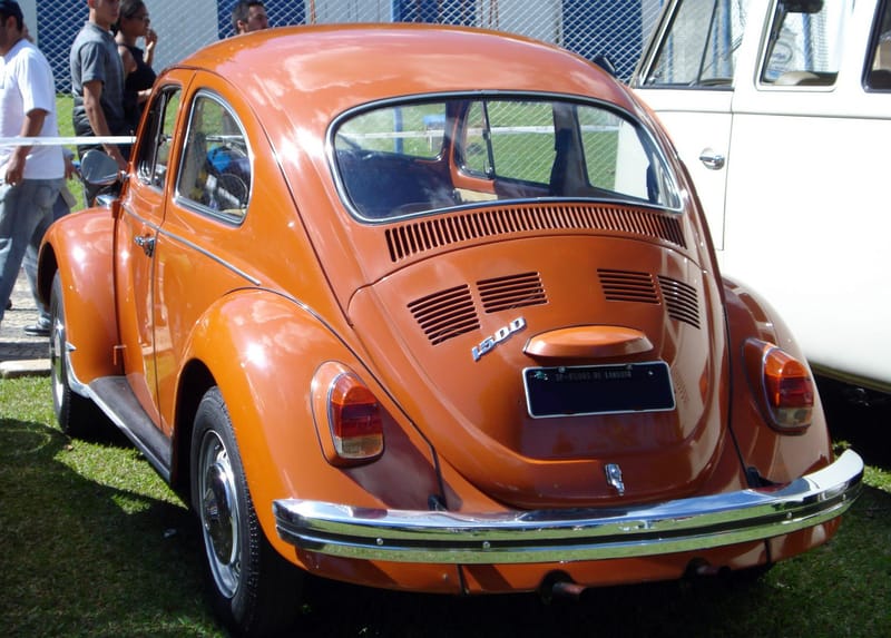 Fusca 1500 laranja visto de trás em gramado de exposição, com o emblema 1500 na tampa do motor, para-choque cromado de lâmina única e uma Kombi branca estacionada ao lado