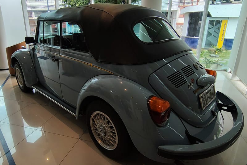 Fusca conversível azul-acinzentado com capota de lona preta fechada, visto de trás e de lado, exposto em salão envidraçado de piso claro