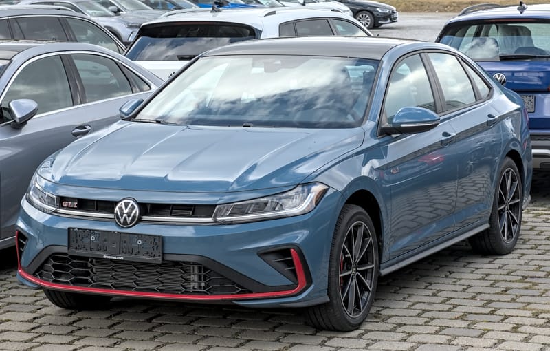 Jetta GLI azul visto de frente e de lado num pátio de carros, com faróis unidos por uma faixa horizontal, emblema GLI vermelho na grade, grade inferior em colmeia e um friso vermelho contornando o para-choque