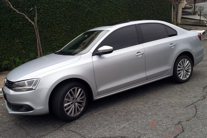 Jetta prata de quatro portas estacionado numa rua em subida, visto de frente e de lado, com rodas de liga de raios finos, teto solar e uma parede coberta de trepadeira ao fundo