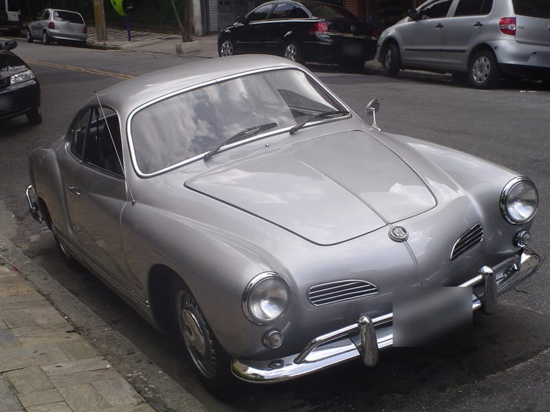 Karmann-Ghia cupê prata estacionado numa rua de São Paulo, com faróis redondos, duas entradas de ar cromadas no bico e para-choque com garras