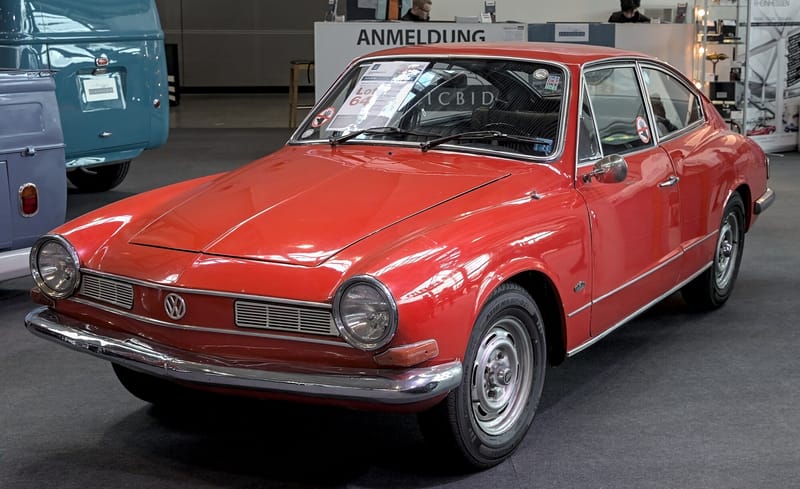 Karmann-Ghia TC vermelho visto de frente e de lado, com faróis redondos, duas grades de frisos horizontais e o emblema VW no centro do bico, exposto num salão de carros antigos