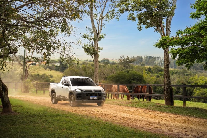 Picape Volkswagen Tukan branca de cabine simples, com para-choque e grade pretos, rodando de frente numa estrada de terra entre árvores; atrás de uma cerca de madeira, cavalos pastam num campo