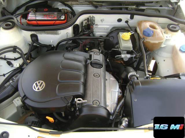 Cofre de um Volkswagen Gol de segunda geração com o motor 1.6 montado no sentido longitudinal, coberto por uma capa plástica preta com o logotipo VW, e um selo gráfico 1.6 Mi no canto da foto