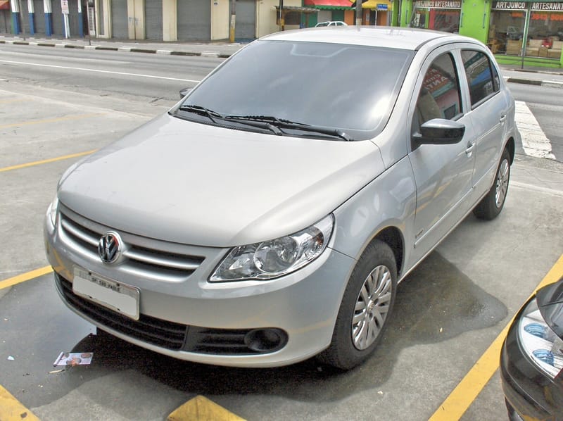 Volkswagen Gol prata de quatro portas parado numa vaga demarcada em amarelo, fotografado pela dianteira e pela lateral, com grade de frisos horizontais prateados, faróis de lente lisa e rodas com calotas