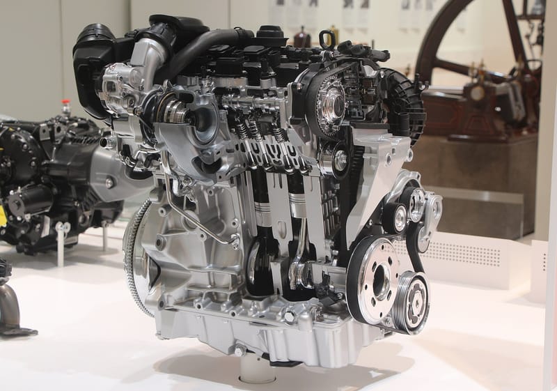 Motor exposto em corte num museu, com bloco e cabeçote de alumínio abertos mostrando pistões, bielas e molas de válvula; à direita, a engrenagem do comando de válvulas envolvida por uma correia dentada preta que desce rumo ao virabrequim