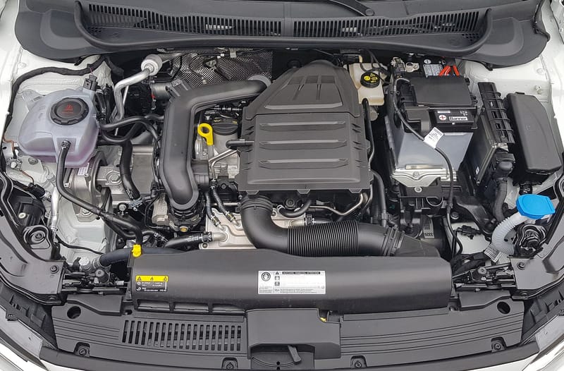 Cofre de um Volkswagen Polo branco visto de cima, com o motor de três cilindros sob uma capa plástica preta, duto de admissão sanfonado à frente, vareta de óleo amarela à esquerda da capa, reservatório de expansão à esquerda e bateria à direita