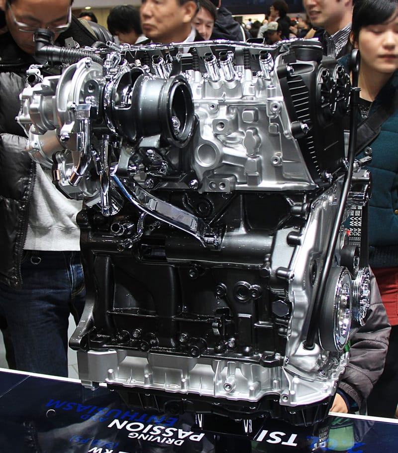 Motor 2.0 TSI exposto num estande, visto pelo lado do turbocompressor, com o bloco escuro, o cabeçote prateado e a tampa da corrente de comando à direita; visitantes ao fundo