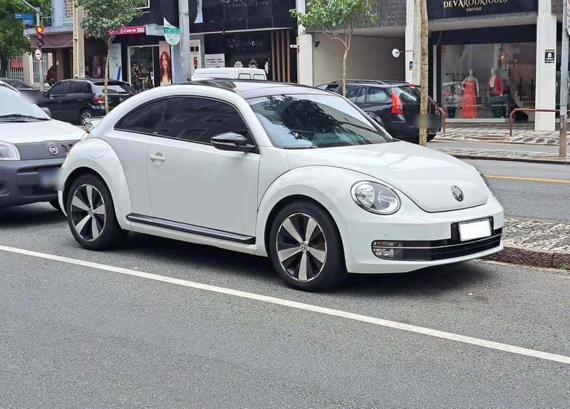 Fusca de terceira geração branco, visto de frente e de lado, estacionado numa rua comercial, com faróis redondos, teto escuro e rodas de liga leve de cinco raios curvos, em preto e prata