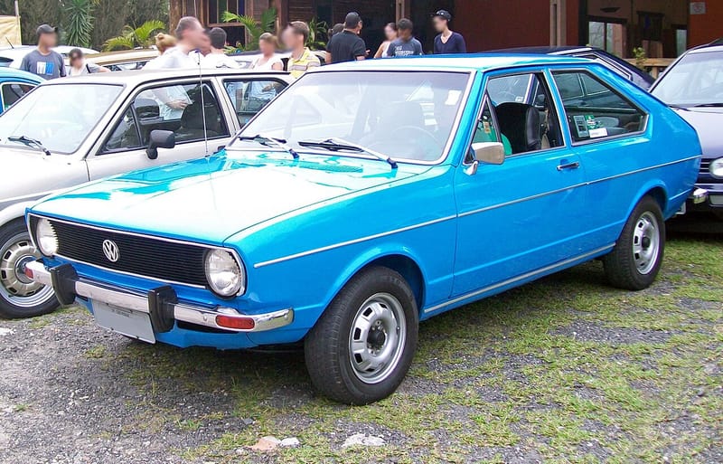 Passat azul de duas portas visto de frente e de lado num encontro de carros antigos, com faróis redondos, grade preta e para-choque cromado com protetores de borracha