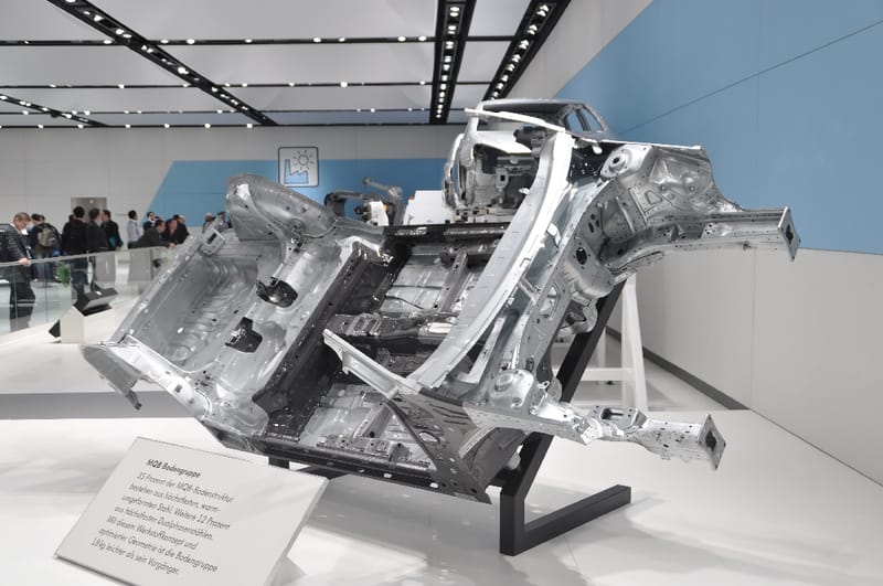 Estrutura de assoalho de automóvel em aço sem pintura, montada inclinada sobre um suporte preto num estande de feira, com uma placa explicativa em alemão em primeiro plano e visitantes ao fundo