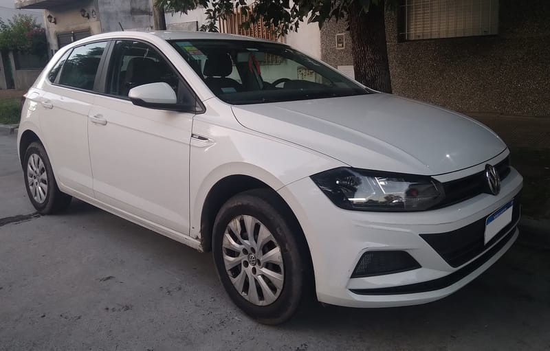 Volkswagen Polo branco de cinco portas visto de frente e de lado, estacionado numa rua de bairro, com calotas prateadas, faróis estreitos e grade fina ligando os faróis