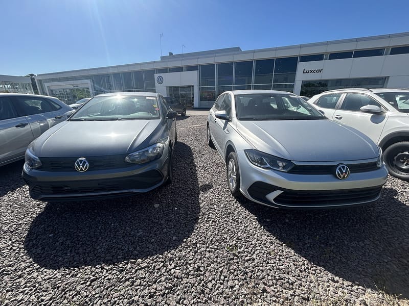 Dois Volkswagen Polo estacionados lado a lado sobre cascalho, de frente, diante de uma concessionária: à esquerda um cinza-escuro com grade superior e entrada de ar inferior pretas, à direita um prata com um filete na cor da carroceria ligando os faróis e grade inferior mais larga