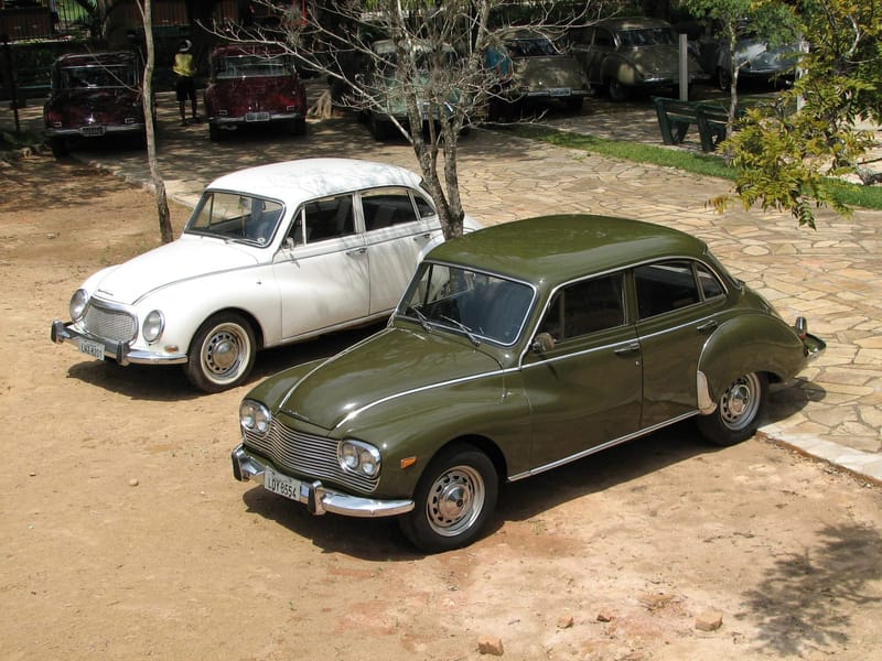 Dois sedãs DKW-Vemag Belcar estacionados sobre terra batida: à frente, um verde-oliva com quatro faróis e grade larga de frisos horizontais; atrás, um branco com dois faróis redondos e grade oval