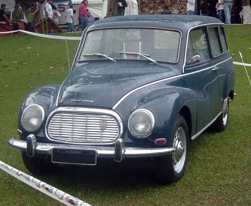 Perua DKW-Vemag Vemaguet azul-escura vista de frente e de lado, com grade oval cromada, faróis redondos e a inscrição DKW-Vemag no capô, estacionada na grama molhada de um encontro de carros antigos
