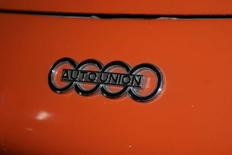Emblema cromado de quatro anéis entrelaçados, cortado por uma faixa com a inscrição AUTO UNION, aplicado na carroceria laranja de um carro