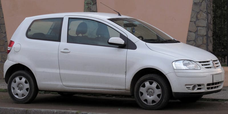 Volkswagen Fox branco de duas portas visto de lado, estacionado junto a um muro, com faróis de desenho arredondado, calotas nas rodas de aço e para-choques na cor da carroceria