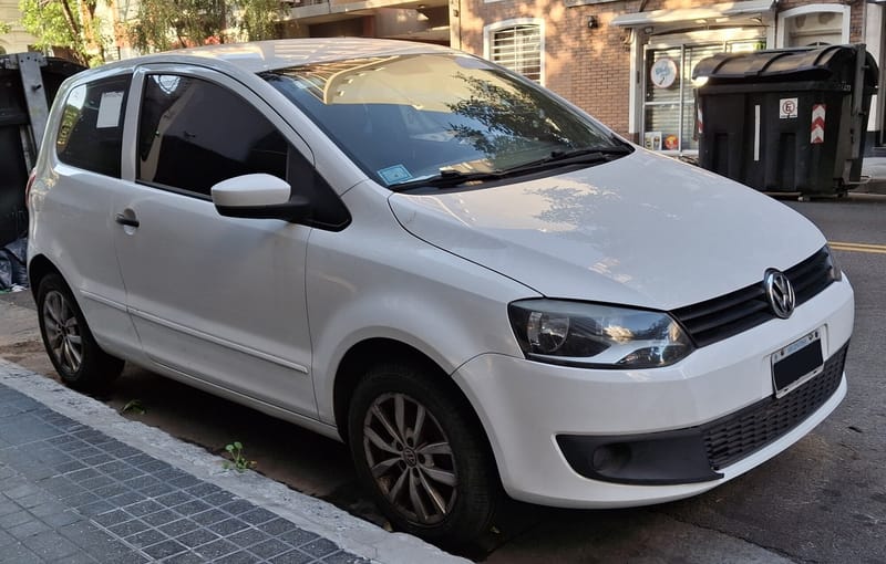 Volkswagen Fox branco de duas portas visto de frente e de lado junto à calçada, com grade preta de frisos horizontais entre faróis alongados e rodas de liga leve