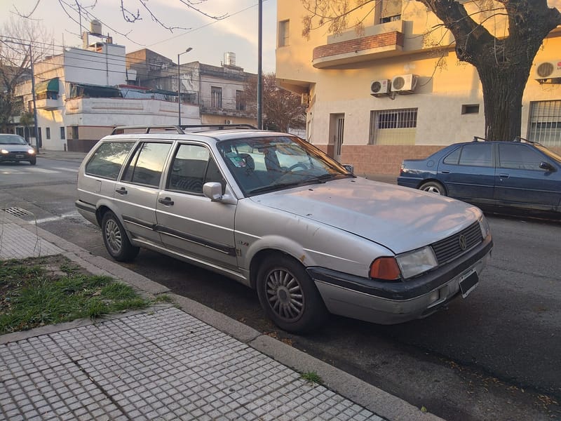 Perua Quantum prata com quatro portas laterais e bagageiro no teto, estacionada junto à calçada, vista de frente e de lado