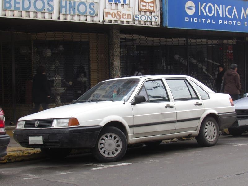 Santana branco de quatro portas estacionado numa rua comercial, com frente em cunha, faróis com piscas laranja nas pontas, para-choques pretos e pequena janela triangular na porta dianteira