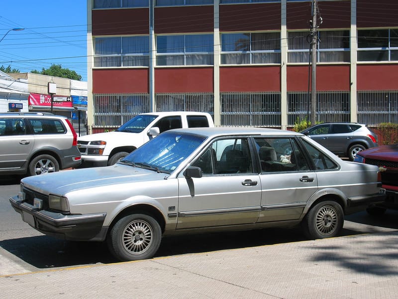 Santana prata de quatro portas estacionado numa rua, visto de frente e de lado, com faróis retangulares, para-choques pretos estreitos e rodas raiadas