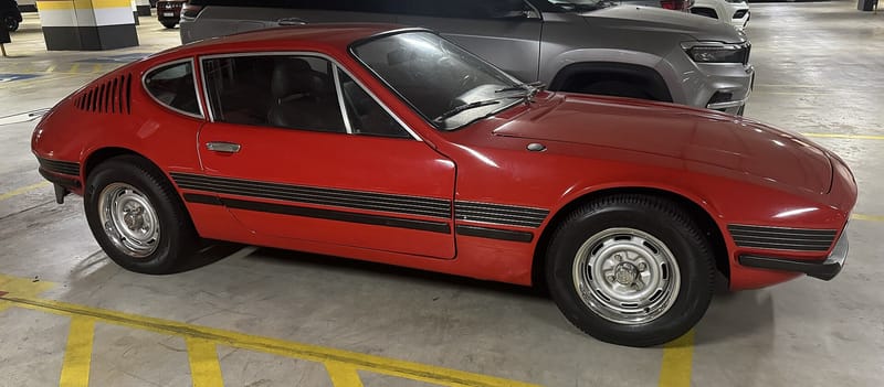 VW SP2 vermelho visto de lado numa garagem, com faixas pretas na lateral, entradas de ar atrás da janela traseira e rodas de aço