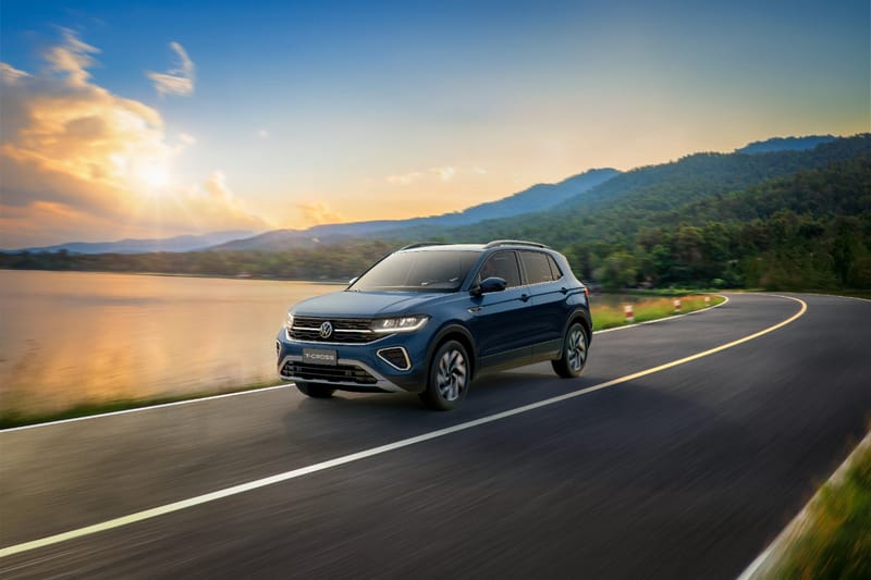 Volkswagen T-Cross azul-escuro rodando numa estrada à beira de um lago, ao pôr do sol, em ângulo de três quartos pela dianteira, com os faróis de LED acesos, filete de luz na grade e molduras iluminadas nas entradas de ar dos cantos do para-choque