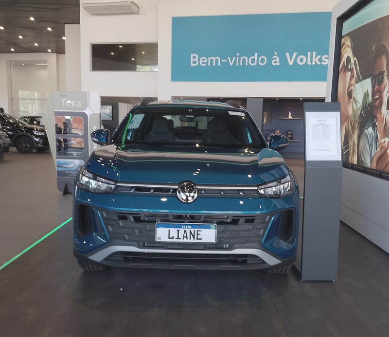 Volkswagen Tera azul visto de frente no salão de uma concessionária, com faróis de LED estreitos ligados por um filete cromado, grade inferior larga em preto e placa decorativa com o nome Liane