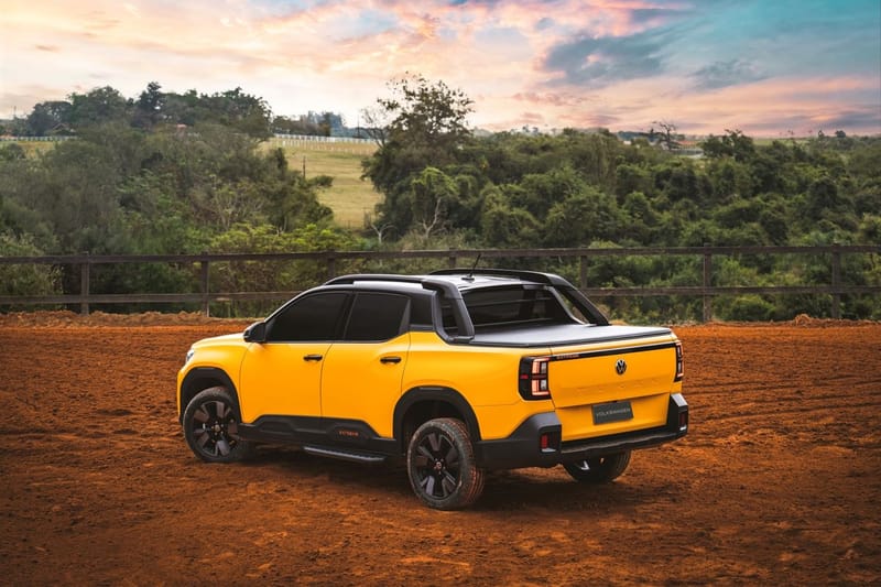 Picape Volkswagen Tukan amarela de cabine dupla, vista de trás e de lado sobre chão de terra vermelha, com o nome Tukan estampado em relevo na tampa da caçamba, lanternas de LED, santantônio preto e emblemas Extreme; ao fundo, uma cerca de madeira e mata